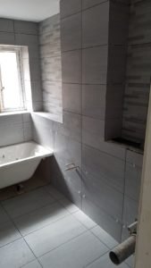 Bathroom under renovation with gray tile walls and floor, a white bathtub along the left wall, a window above it, and an inset tiled niche in the wall; exposed pipes and spacers visible indicating ongoing plumbing work.
