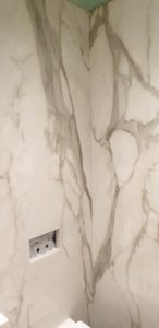 Marble walls with gray veins meeting at a vertical seam, showing a recessed electrical outlet box in the lower left corner of the wall (bathroom area).