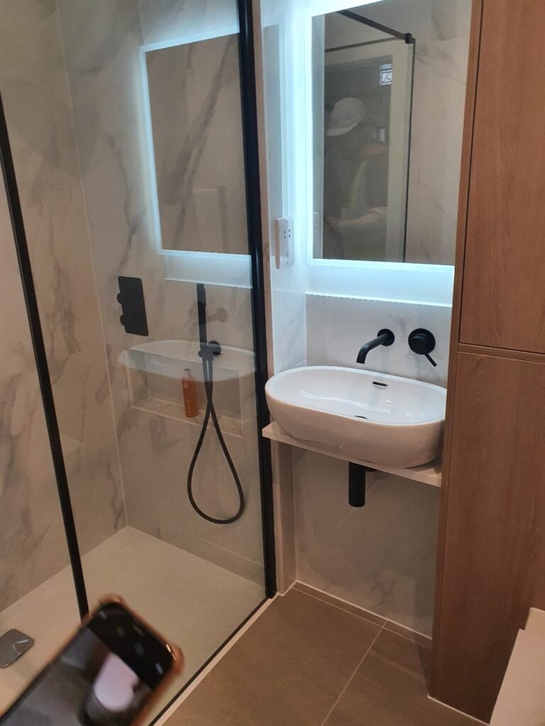 Modern bathroom with a glass shower door, marble walls, and a white wall-mounted sink with black fixtures.