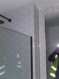 Bathroom with white hexagonal tiles, glass shower door, and a mirror reflecting a person in a high-visibility vest.
