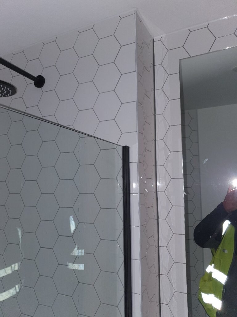 Bathroom shower with white hexagonal tiles and a glass door; reflection shows a person in a high-visibility vest taking the photo.