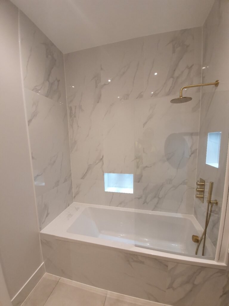 Marble-tiled shower with a glass panel, brass rain showerhead, and a handheld shower fixture; built-in recessed niches glow with blue light.