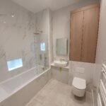Modern marble-tiled bathroom with a glass shower screen, built-in bathtub, wall-mounted sink, and a wooden wall cabinet above a wall-mounted toilet in a light grey palette.