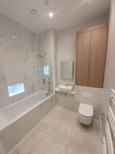 Modern marble-tiled bathroom with a glass shower screen, built-in bathtub, wall-mounted sink, and a wooden wall cabinet above a wall-mounted toilet in a light grey palette.