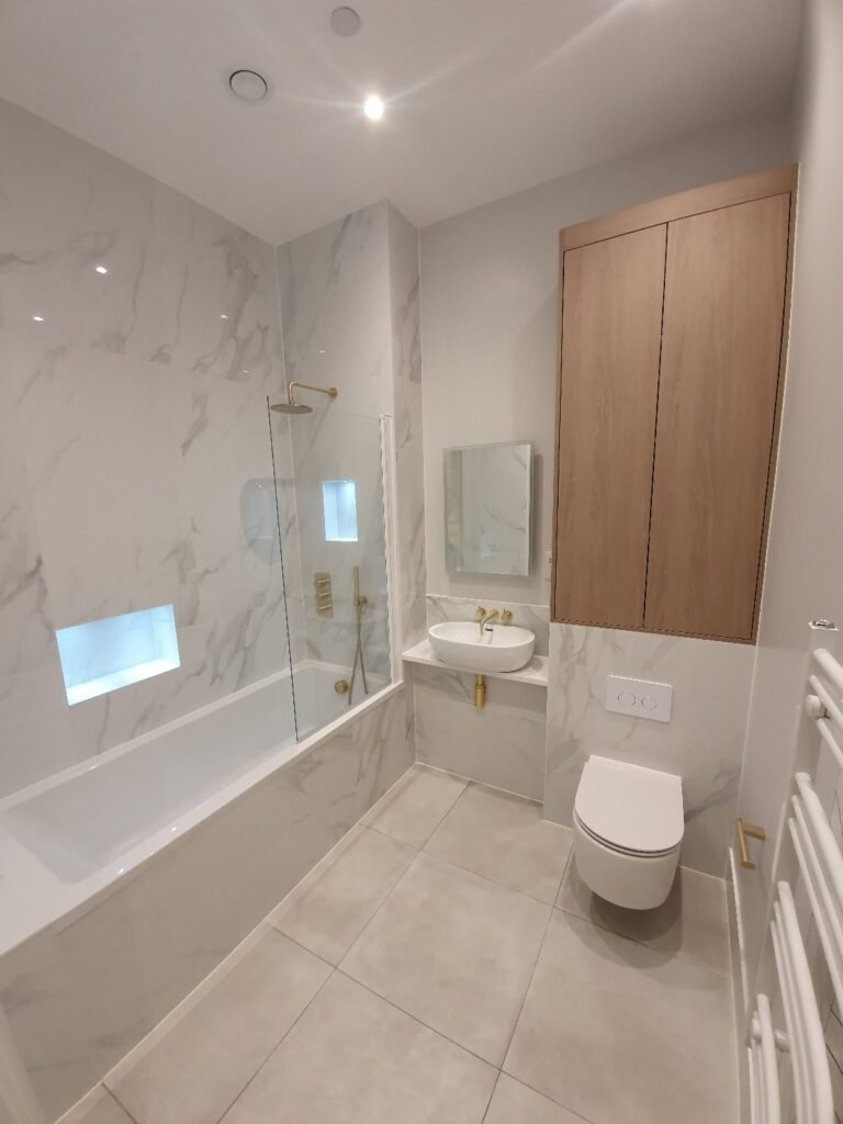 Modern marble-tiled bathroom with a glass shower screen, built-in bathtub, wall-mounted sink, and a wooden wall cabinet above a wall-mounted toilet in a light grey palette.