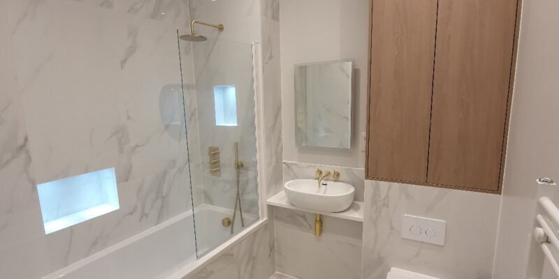 Modern marble-tiled bathroom with a glass shower screen, built-in bathtub, wall-mounted sink, and a wooden wall cabinet above a wall-mounted toilet in a light grey palette.