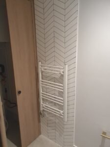 White wall-mounted towel radiator on a chevron-tiled corner, adjacent to a wooden door and white wall.