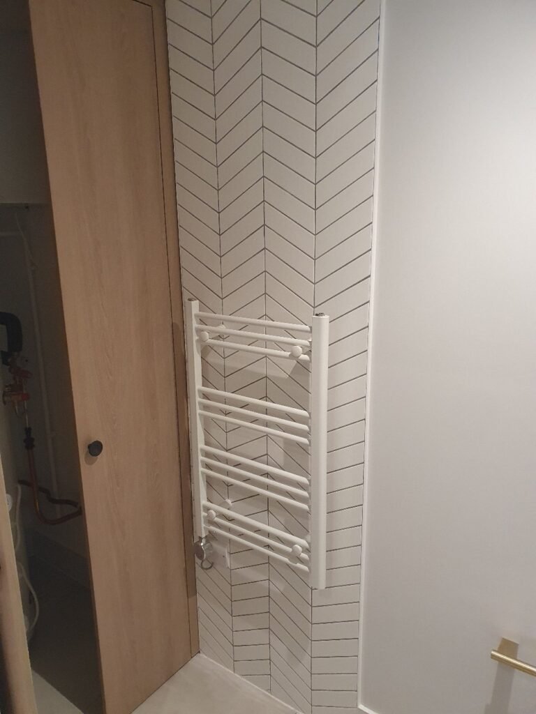 White towel radiator mounted on a chevron-tiled wall beside a wooden door in a bathroom.