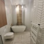 Modern bathroom with a white freestanding tub centered beside a tall stone pillar and beige tile floor; toilet and wooden cabinet visible.