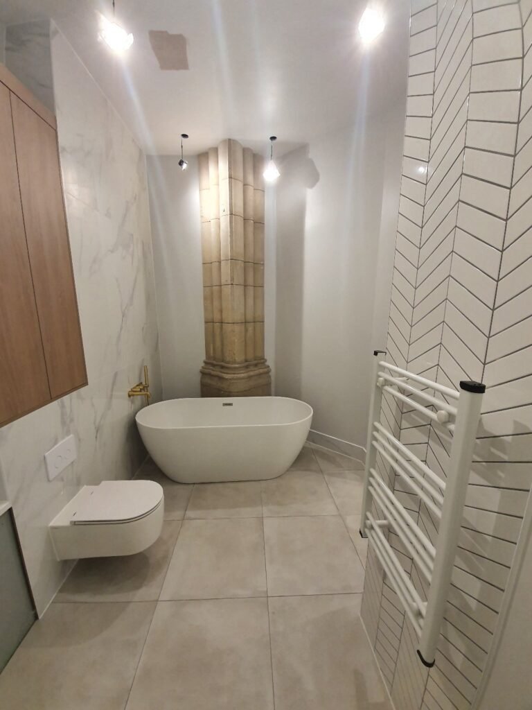 Modern bathroom with a white freestanding tub beside a stone column, large light tiles, and a wall-mounted toilet nearby.