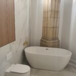 Modern bathroom with a freestanding white tub beside a large stone column and marble wall panels.