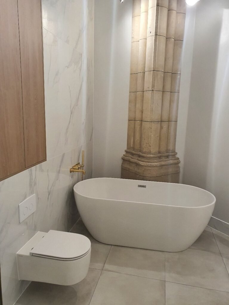 Modern bathroom with a freestanding white tub, wall-mounted toilet, marble walls, and a large stone column in the corner.